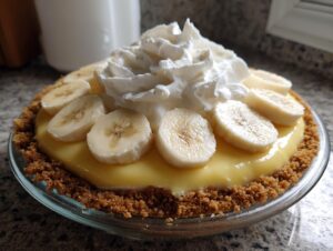 A close-up of a homemade banana cream pie featuring a graham cracker crust, yellow filling, sliced bananas, and a large dollop of whipped cream.