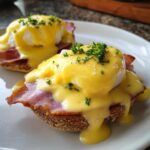 Two servings of classic eggs benedict featuring poached eggs, ham, and rich hollandaise sauce, garnished with parsley.