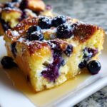 A close-up of a moist slice of Blueberry French Toast Casserole topped with fresh blueberries and powdered sugar, sitting in syrup.