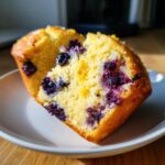 A bright yellow blueberry protein muffin cut in half, showing moist crumb and burst blueberries, sprinkled with sugar.