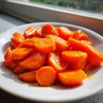 A close-up of bright orange, shiny glazed carrots served on a white plate near a window.