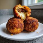 Three golden-brown, deep-fried rice balls stacked on a white plate, one cut open showing melted cheese and meat filling.