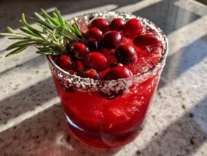 A vibrant red cranberry pomegranate margarita garnished with fresh cranberries and a rosemary sprig, featuring a salted rim.