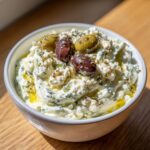 Close-up of a bowl of creamy dirty martini dip, topped with green and Kalamata olives and herbs.