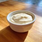 A small white bowl filled with thick, creamy homemade garlic sauce sitting on a wooden cutting board.