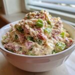 A close-up of a white bowl filled with creamy ham salad mixed with visible chunks of pink ham and bright green celery.