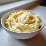 A close-up of creamy slow cooker mashed potatoes topped with melting butter and black pepper in a white bowl.