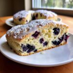 Close-up of a moist slice of blueberry scones, topped with powdered sugar and a white glaze drizzle.