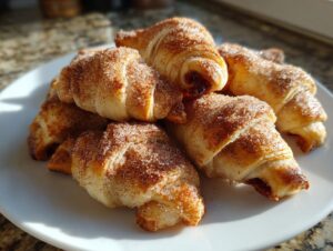 A pile of golden brown, crescent-shaped Rugelach pastries dusted heavily with cinnamon sugar on a white plate.