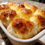 Close-up of freshly baked, golden brown homemade dinner rolls topped with melted butter or glaze in a white ceramic dish.