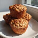 Three golden brown banana oatmeal muffins stacked on a white plate, catching bright sunlight.