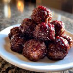 A small pile of glistening grape jelly meatballs coated in a thick, dark red glaze, served on a white plate.
