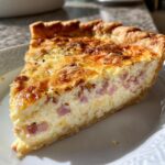 A golden-brown slice of ham and cheese quiche on a white plate, showing creamy filling with diced ham.