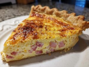 Close-up of a thick slice of ham and cheese quiche showing the golden-brown top and chunks of ham in the creamy filling.