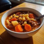 A close-up of a white bowl filled with rich hamburger soup, featuring ground beef, rotini pasta, potatoes, and bright orange carrots.