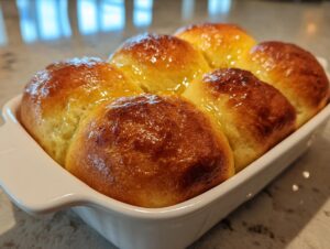Six golden brown homemade dinner rolls baked together in a white dish, glistening with a buttery glaze.