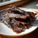 A pile of dark, seasoned strips of homemade venison jerky resting on a white plate.