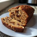 A moist slice of date nut bread showing visible dates and walnuts, resting next to the main loaf on a white plate.