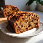 Two thick slices of moist fruit cake, packed with raisins and nuts, served on a white plate.