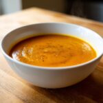 Close-up of a white bowl filled with vibrant orange roasted butternut squash soup, topped with pepper.