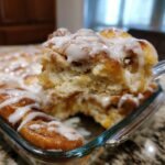 A close-up of a fork lifting a gooey, frosted piece of cinnamon roll casserole from a glass baking dish.