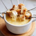 Close-up of a white bowl filled with smooth cheese fondue, featuring toasted bread cubes being dipped on skewers.
