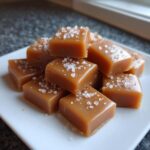 A close-up of soft, square caramels sprinkled with flaky sea salt, piled on a white dish.
