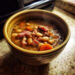 Close-up of a steaming bowl of hearty Ham and Bean Soup, featuring white beans, chunks of ham, and sliced carrots.
