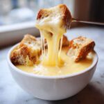 A piece of toasted bread is lifted from a bowl of creamy cheese fondue, showing long, irresistible cheese pulls.