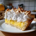 A close-up slice of lemon meringue pie showing bright yellow curd, flaky crust, and tall, torched meringue.