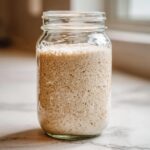 A glass jar filled with active, bubbly sourdough starter showing significant aeration and rise.