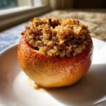 A single, glistening baked apples dessert with a crumbly oat topping served on a white plate.