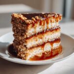 A tall slice of caramel crunch cake with three layers, dripping with caramel sauce and coated in nuts.