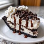 A thick slice of creamy ice cream pie with a dark crust, topped with whipped cream, chocolate sauce, and mini chocolate chips.