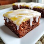A close-up of a rich, moist square of lemon brownies topped with a yellow layer and drizzled with white icing.