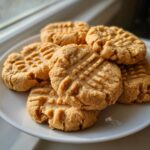 A stack of golden brown peanut butter cookies featuring classic crisscross fork marks, resting on a white plate.