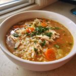 Close-up of a white bowl filled with rich turkey soup, featuring shredded meat, rice, carrots, and fresh parsley garnish.