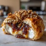 A close-up of a slice of baked brie wrapped in flaky pastry, oozing melted cheese, fig jam, and topped with candied walnuts.