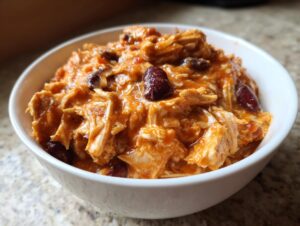 Close-up of shredded chicken and kidney beans coated in a creamy, orange sauce, representing delicious buffalo chicken chili, served in a white bowl.