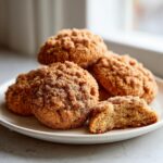 A plate of soft coffee cake cookies generously coated in cinnamon sugar crumb topping, one cookie is broken open.