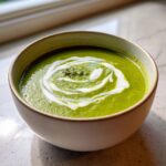 A close-up of a bowl of vibrant green asparagus soup, topped with a swirl of cream and whole asparagus tips.