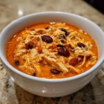 Close-up of a white bowl filled with creamy, orange-red buffalo chicken chili featuring shredded chicken and dark red kidney beans.