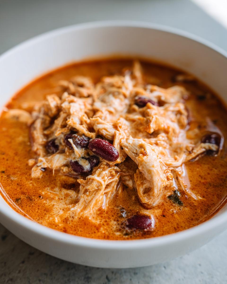A close-up of creamy buffalo chicken chili featuring shredded chicken and dark red kidney beans in a rich orange broth.