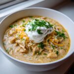 A close-up of a bowl of creamy white chicken chili topped with shredded cheese, sour cream, and fresh parsley.