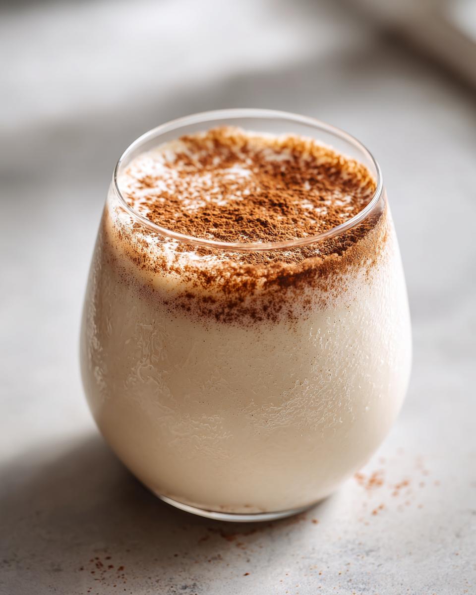 Close-up of a glass filled with creamy, chilled coquito, generously dusted with ground cinnamon on top.