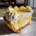 A close-up slice of creamy lemon pie with a graham cracker crust, topped with whipped cream and lemon zest.