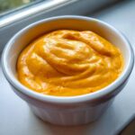 Close-up of thick, vibrant orange nacho cheese sauce swirled in a small white ramekin dish.