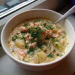 A close-up of a creamy bowl of salmon soup, featuring chunks of pink salmon, potatoes, and topped generously with fresh dill.