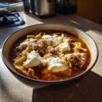 A close-up of a bowl of rich, red crockpot lasagna soup topped with dollops of white ricotta cheese and grated parmesan.