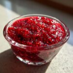 A close-up, vibrant shot of thick, homemade raspberry jam glistening in a small, clear glass bowl.